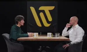 Zwei Personen im Gespräch während einer Podcast-Aufnahme über die digitale Transformation im Unternehmen, sitzend an einem Holztisch mit Mikrofonen vor einem schwarz-gelben Logo-Hintergrund.